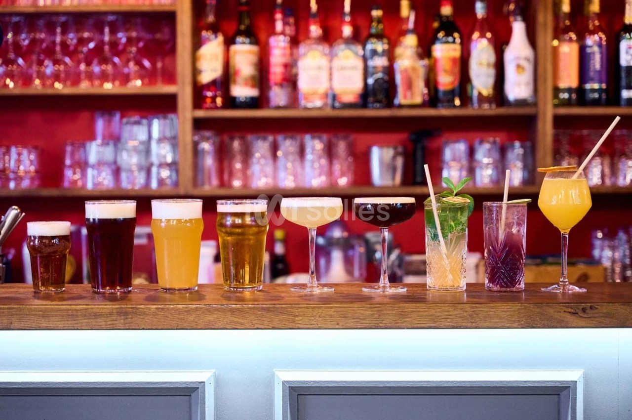 Alignement de verres contenant diverses boissons colorées sur un comptoir de bar, avec des bouteilles en arrière-plan.