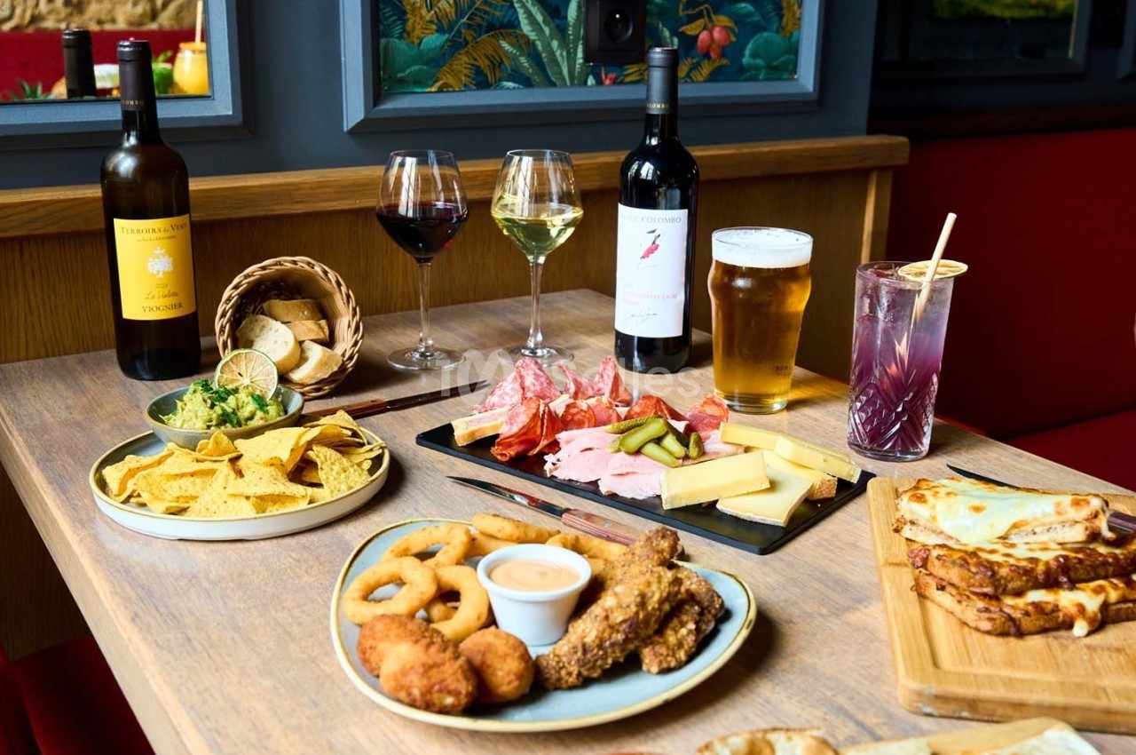 Table garnie de charcuteries, fromages, nachos, beignets, boissons variées et pain, dans un cadre chaleureux.