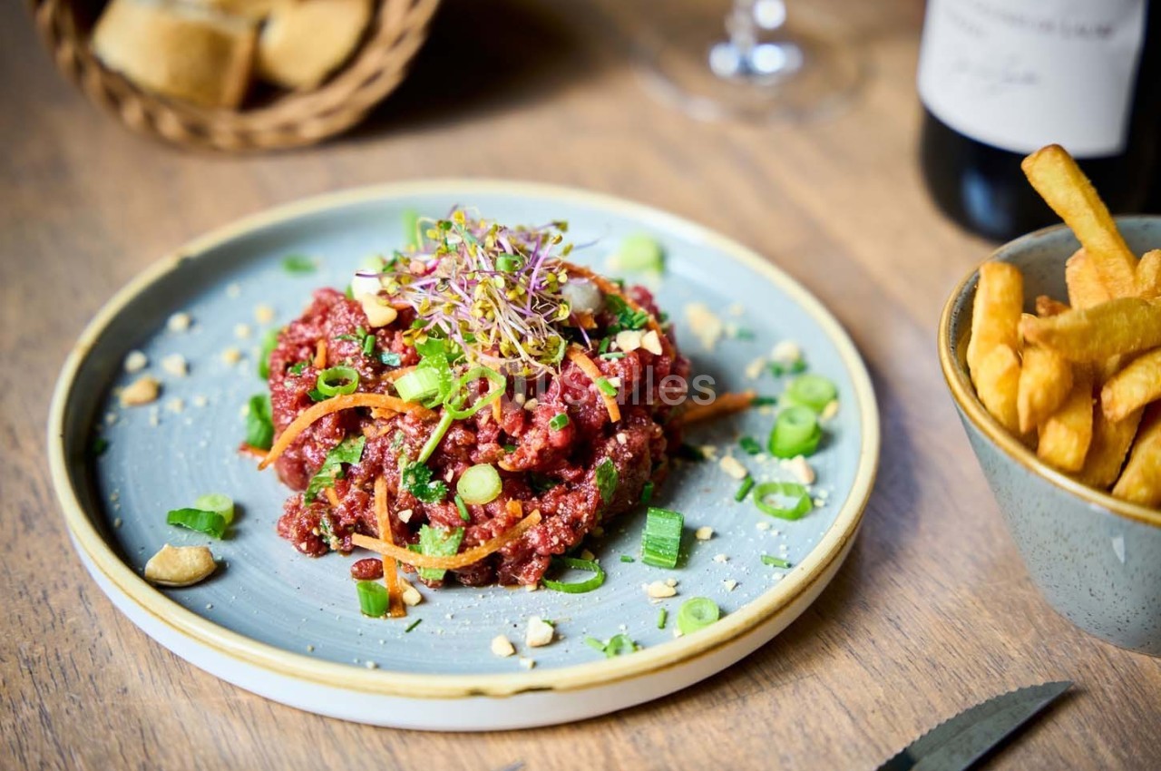 Assiette de tartare de bœuf garni de légumes et herbes, accompagnée de frites et d'un panier de pain en arrière-plan.