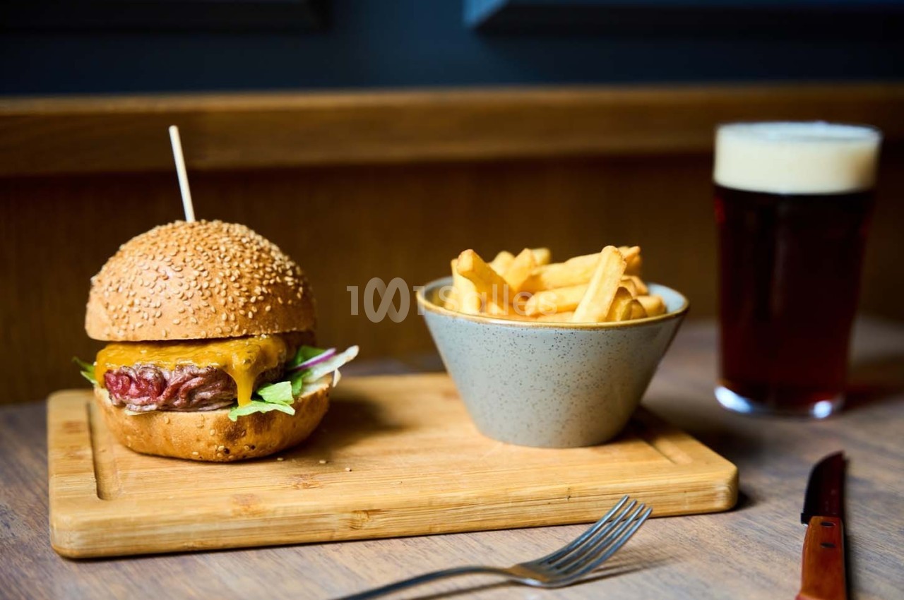 Burger au fromage avec salade, accompagné de frites dans un bol et d'une bière, servi sur une planche en bois.