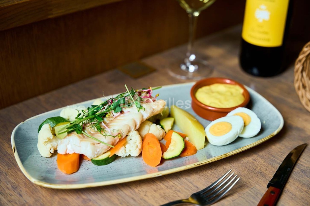 Assiette de poisson accompagné de légumes cuits, œufs durs, sauce jaune et verre de vin en arrière-plan.
