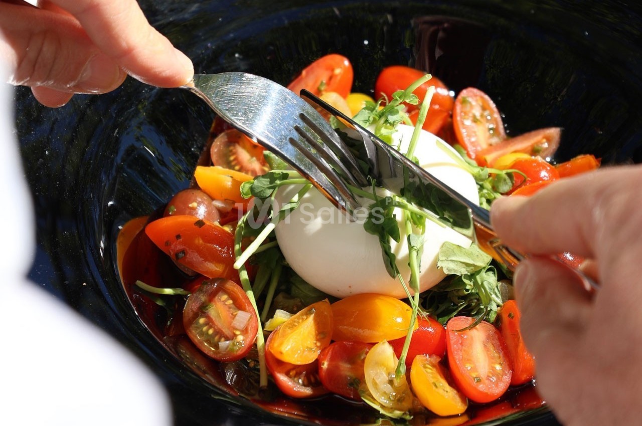 Personne coupant une burrata accompagnée de tomates cerises colorées et de jeunes pousses dans une assiette noire.
