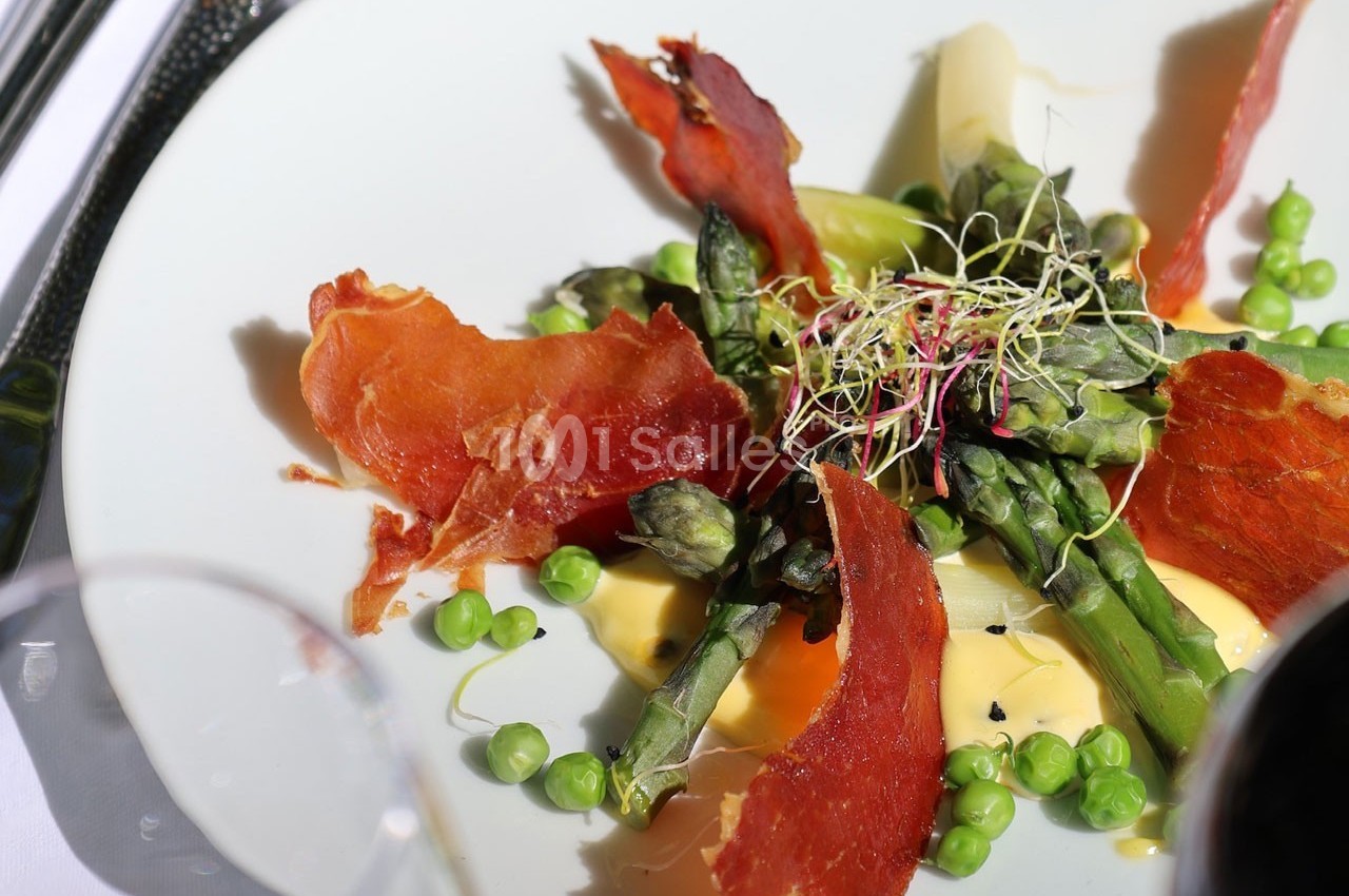 Assiette garnie d'asperges, petits pois, jambon croustillant, germes et sauce crémeuse, présentée sur une table.