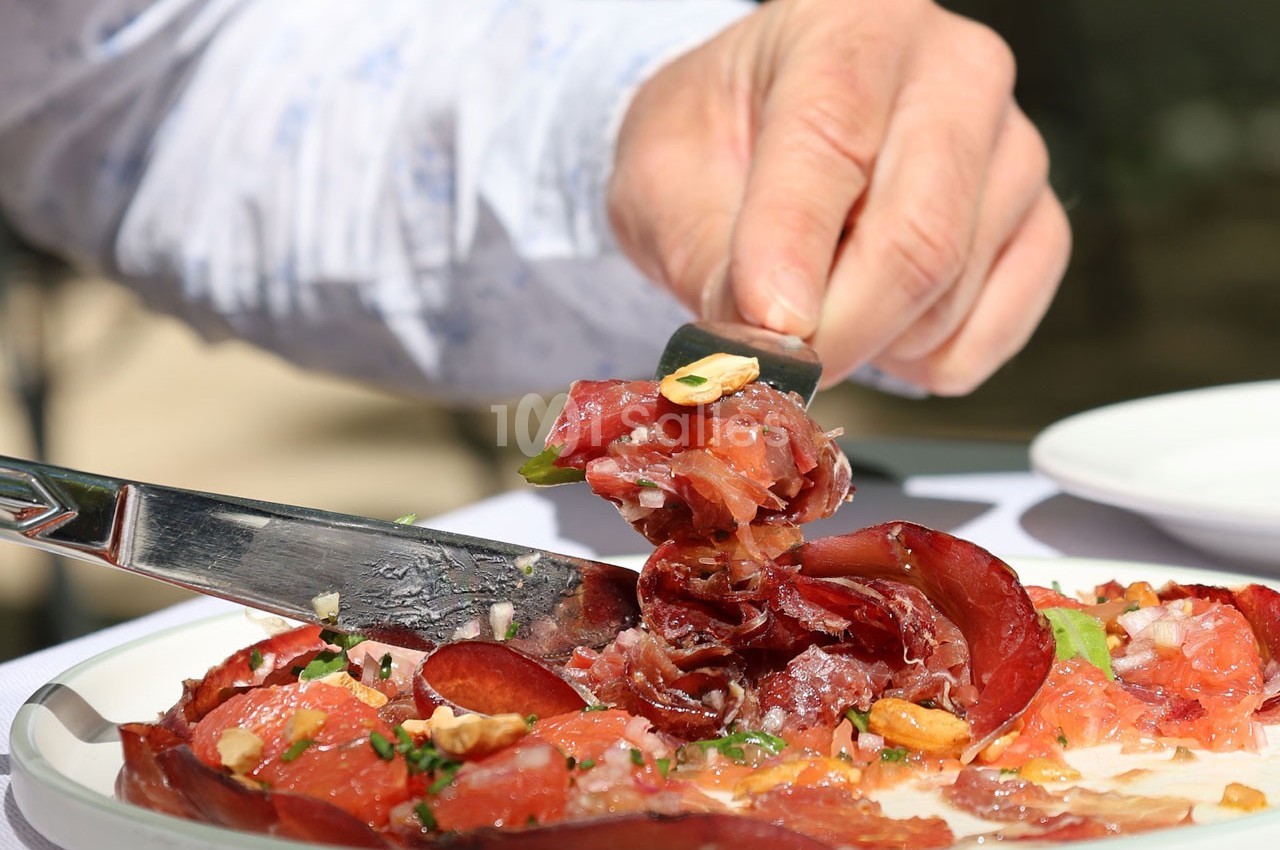Une main utilise un couteau pour servir des tranches de viande séchée garnies de noix et d'herbes sur une assiette.
