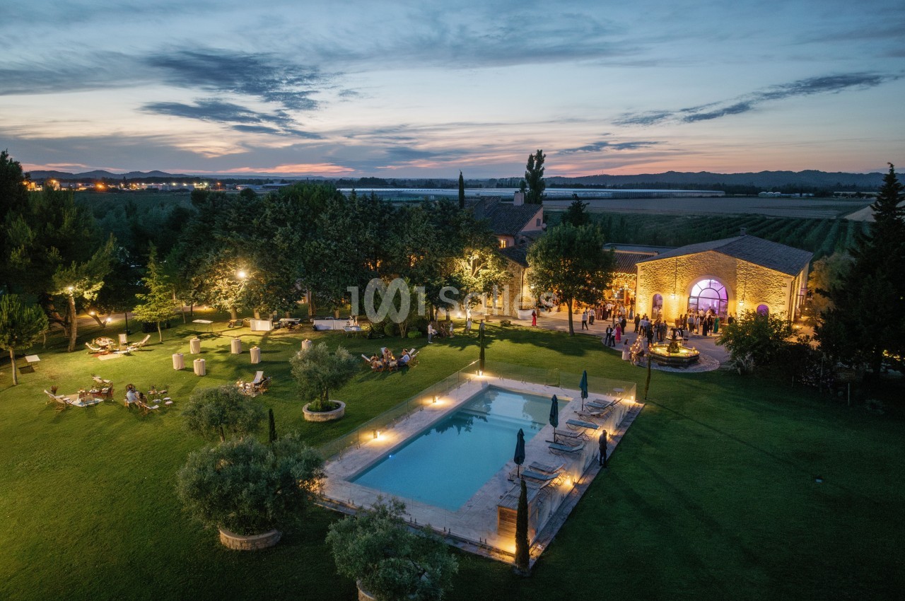 Vue aérienne d'une propriété avec piscine éclairée, jardin verdoyant et bâtiment en pierre au crépuscule.