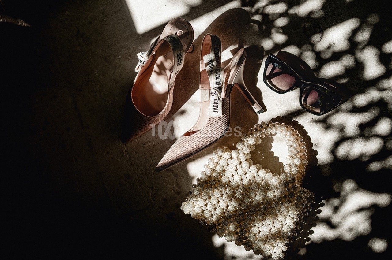 Chaussures à talons, lunettes de soleil et sac en perles posés sur un sol éclairé par une lumière tamisée.