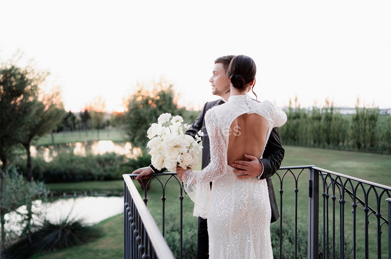 Un couple en tenue de mariage se tient sur un balcon, regardant un paysage verdoyant au coucher du soleil.