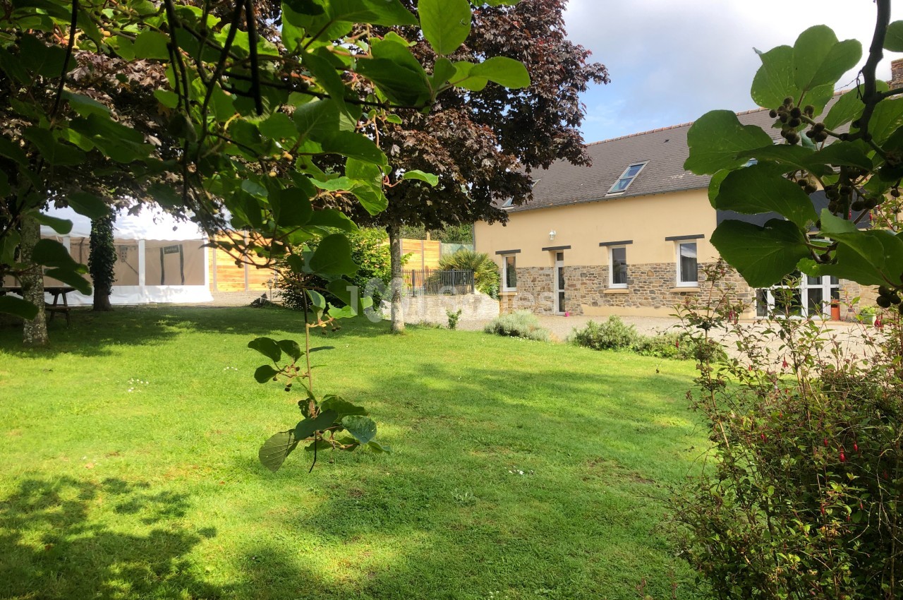 Jardin verdoyant avec pelouse, arbres et arbustes, devant un bâtiment en pierre et crépi jaune sous un ciel partiellement…
