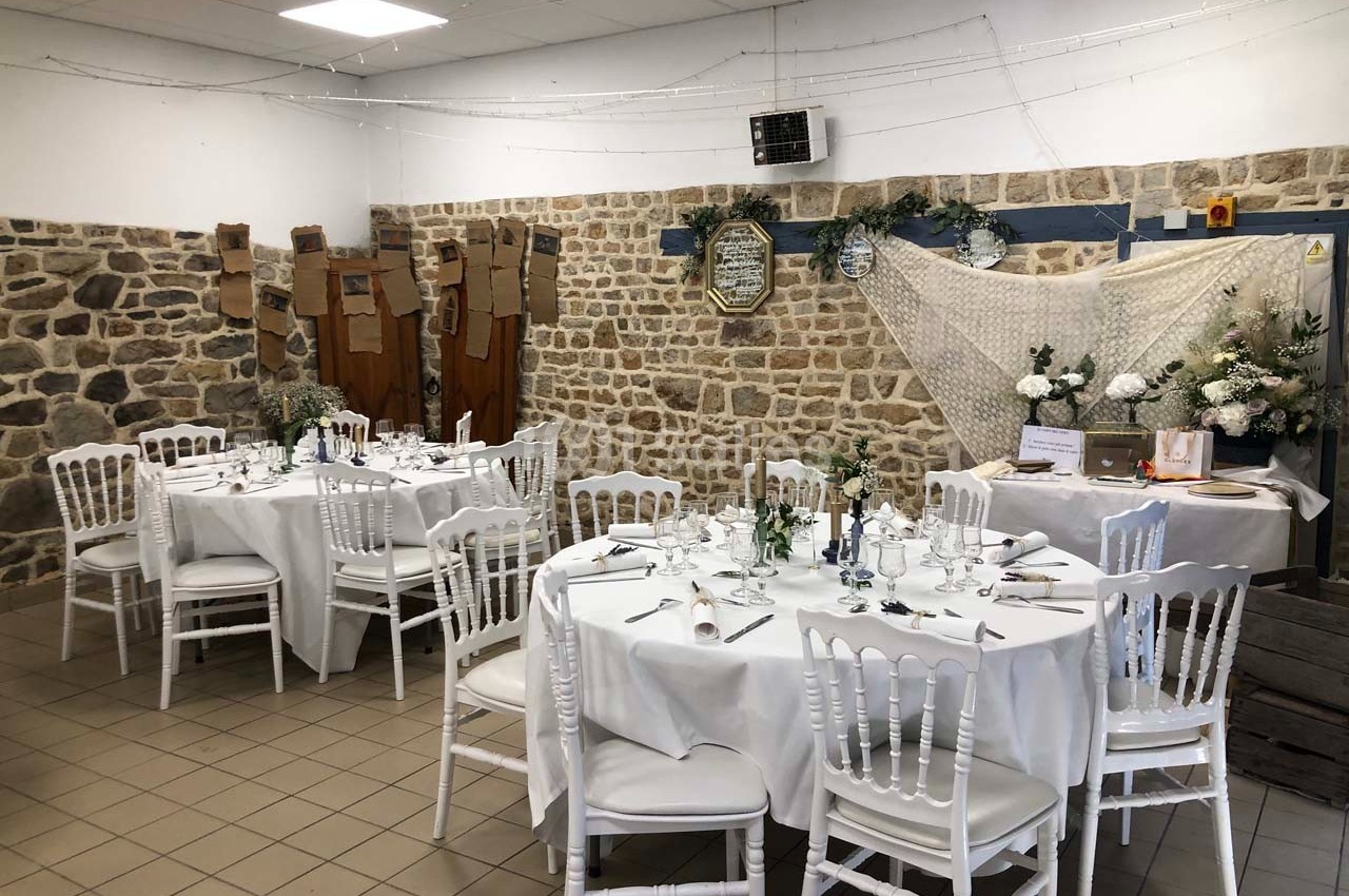 Salle décorée pour un événement avec tables rondes dressées, chaises blanches et murs en pierre apparente.