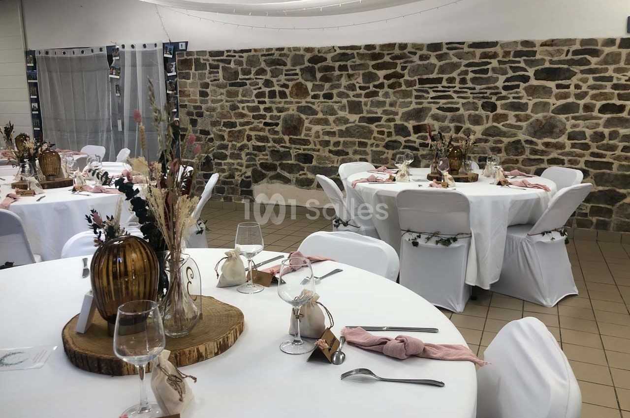 Salle décorée pour un événement avec des tables rondes dressées, nappes blanches et décorations florales rustiques.