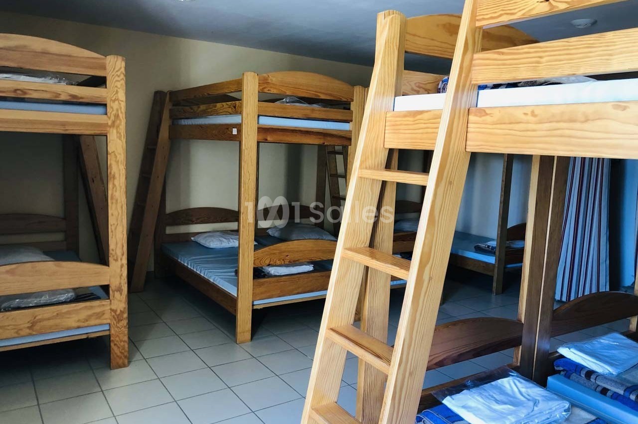 Dortoir avec plusieurs lits superposés en bois, matelas et oreillers, sur un sol carrelé clair.