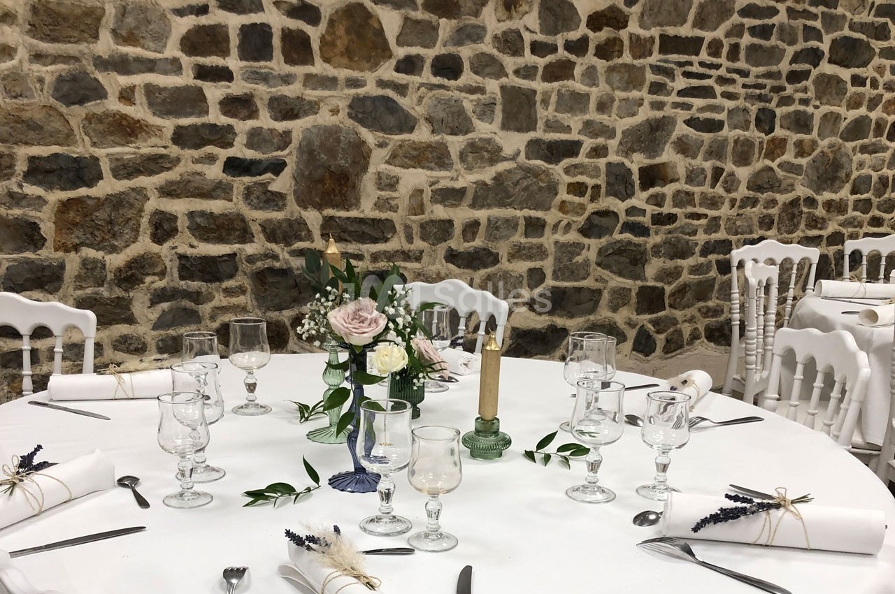 Table ronde décorée pour un événement, avec nappes blanches, verres, couverts, bougie et centre de table floral.