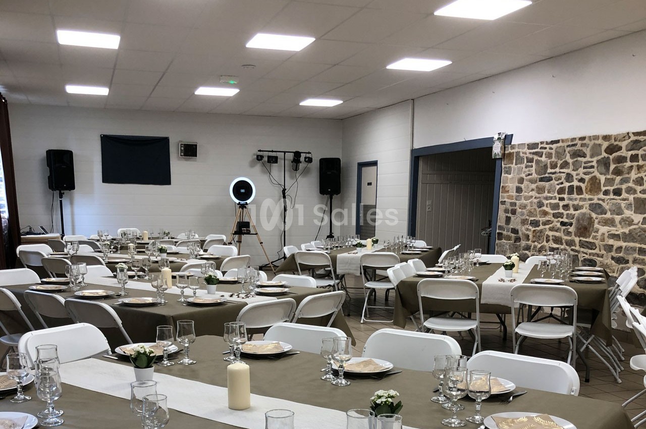 Salle décorée pour un événement avec tables dressées, nappes beige, chaises blanches et équipement audio en arrière-plan.