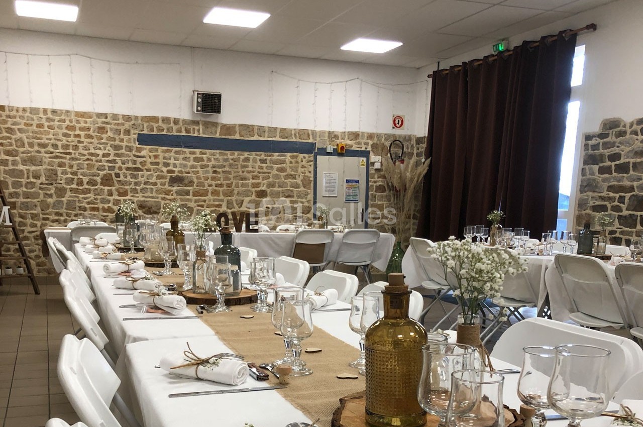 Salle décorée pour un événement avec des tables alignées, nappes blanches, décorations rustiques et murs en pierre.