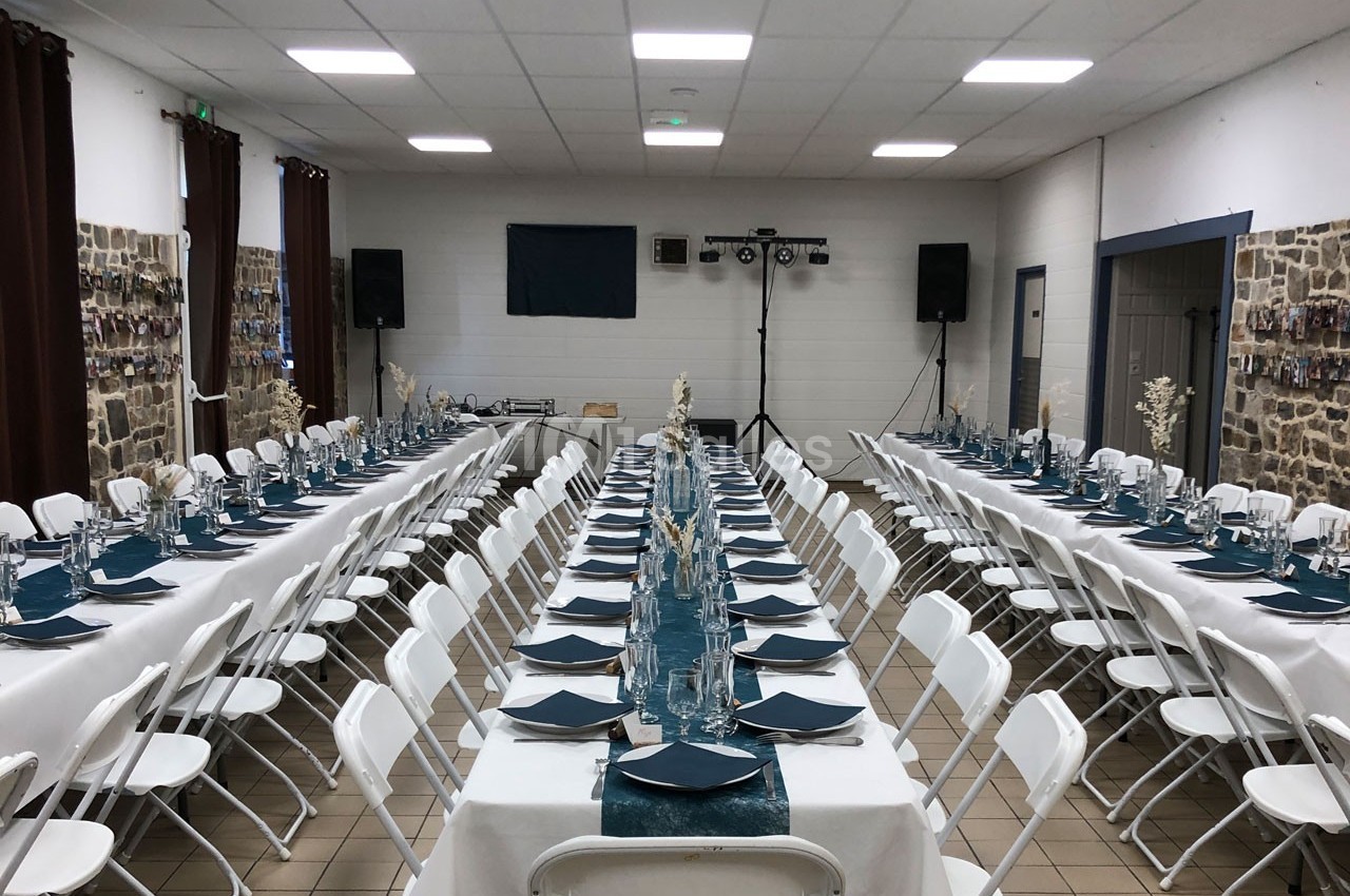 Salle de réception aménagée avec deux longues tables dressées, nappes blanches et décorations bleues, prête pour un…