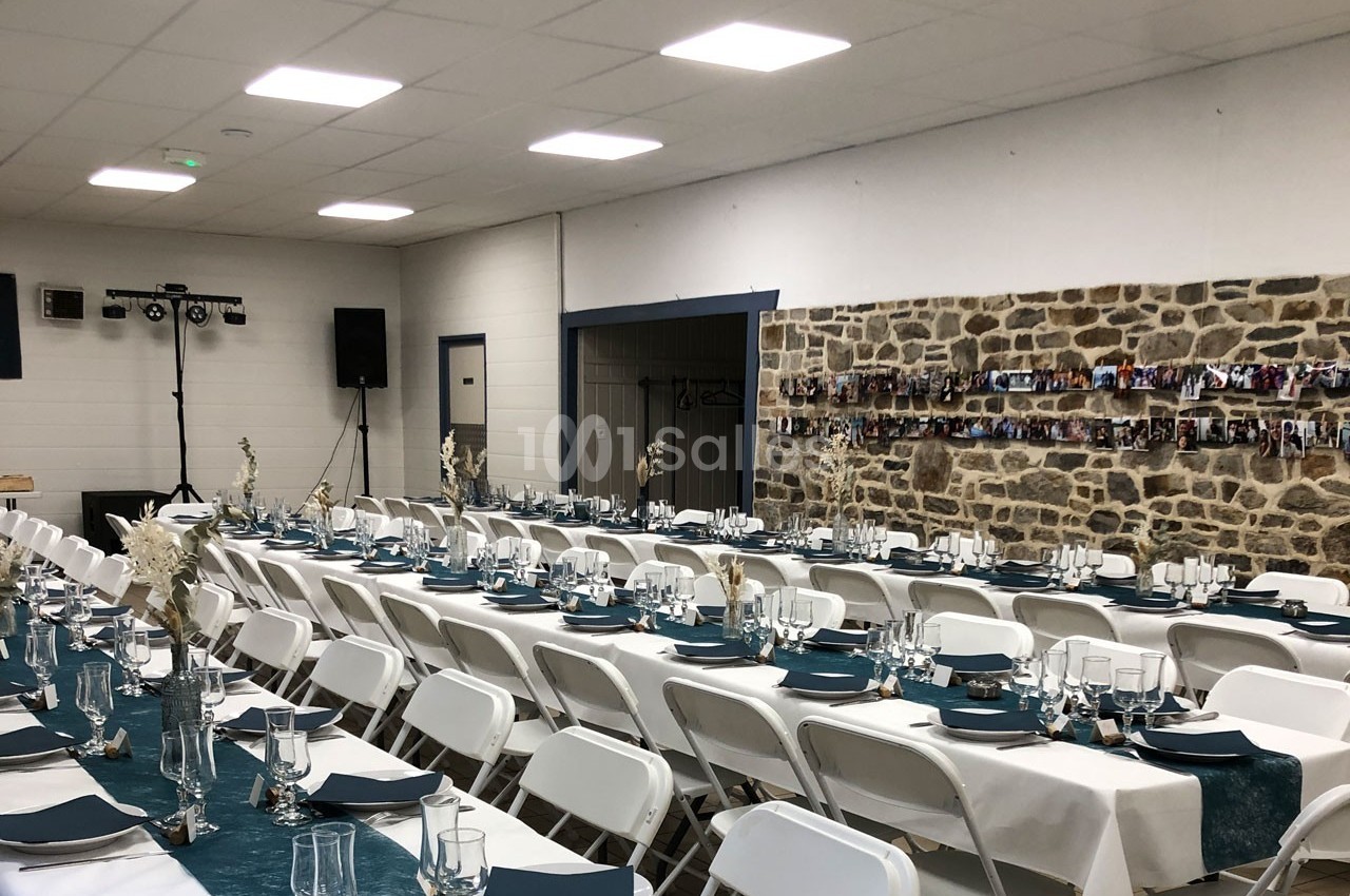 Salle décorée pour un événement avec des tables alignées, nappes blanches, chemins de table bleus et murs en pierre.