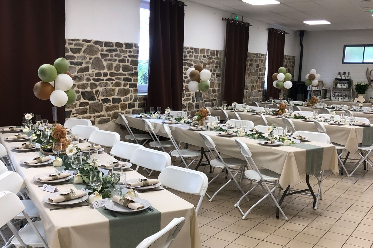 Salle décorée pour un événement, avec tables alignées, nappes beige et verte, ballons et compositions florales.