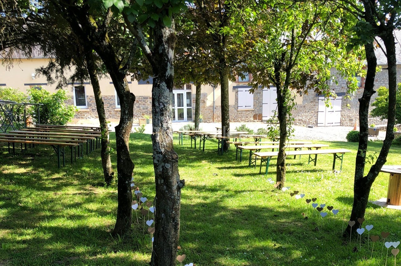 Jardin ombragé avec tables et bancs en bois disposés pour un événement, entouré d'arbres et de décorations en forme de cœurs.