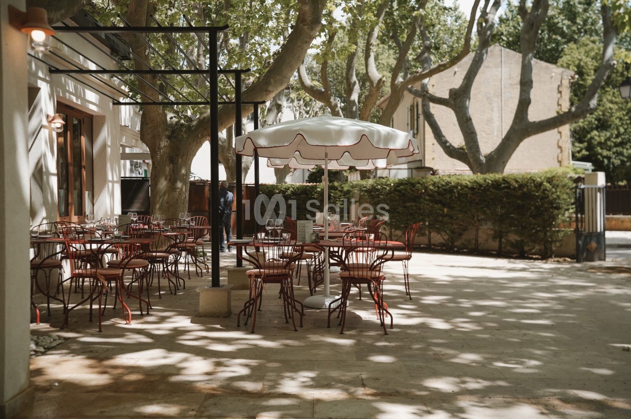 Terrasse d'un café avec des tables et chaises en métal rouge sous un parasol, entourée d'arbres et de lumière naturelle.