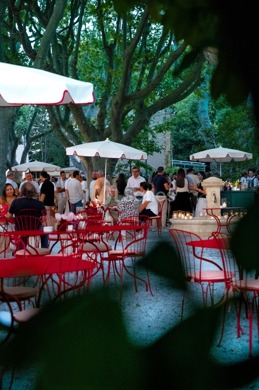 Terrasse animée en soirée avec des tables rouges, des parasols blancs et des convives sous des arbres éclairés.