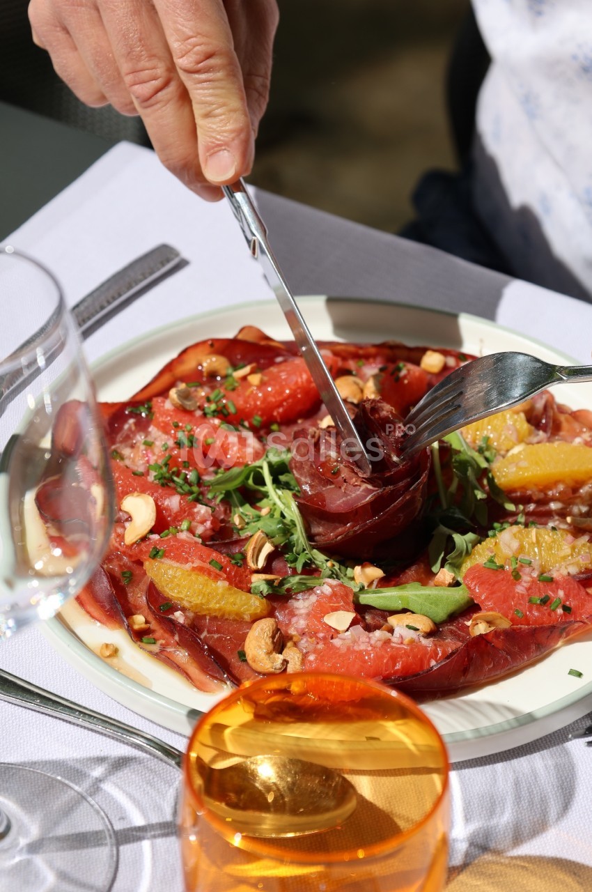 Une assiette de carpaccio de viande avec roquette, agrumes, noix et assaisonnement, une main utilise des couverts.