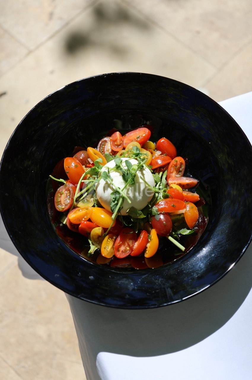 Assiette de burrata garnie de tomates cerises colorées et de roquette, présentée sur une table en extérieur.