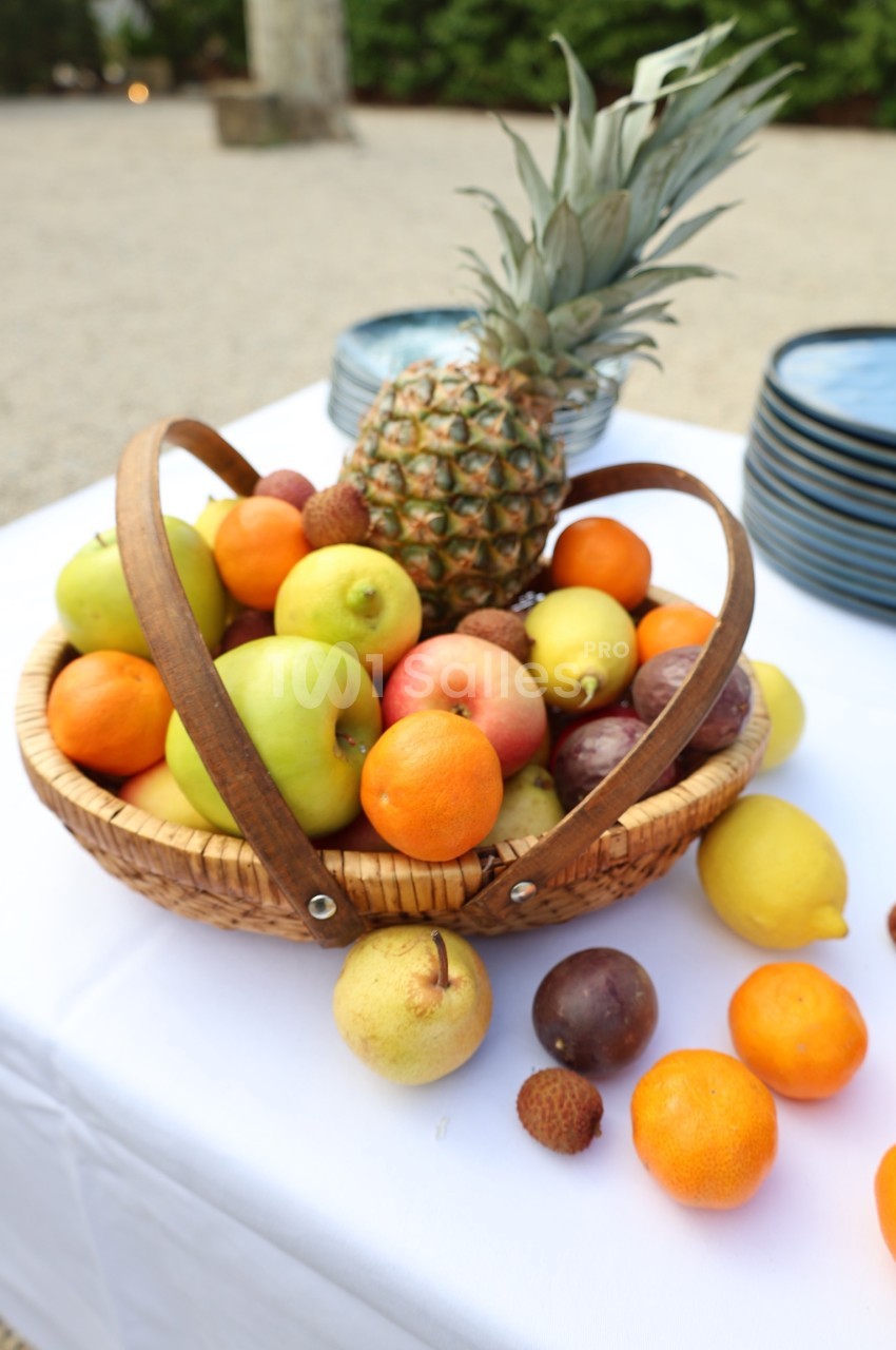 Panier en osier rempli de fruits variés, dont ananas, pommes, oranges et citrons, posé sur une nappe blanche.