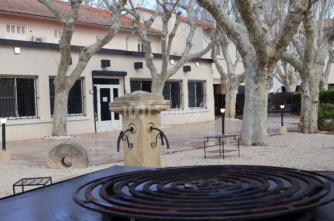 Cour extérieure avec arbres, fontaine en pierre, mobilier de jardin et bâtiment beige aux fenêtres grillagées en arrière…