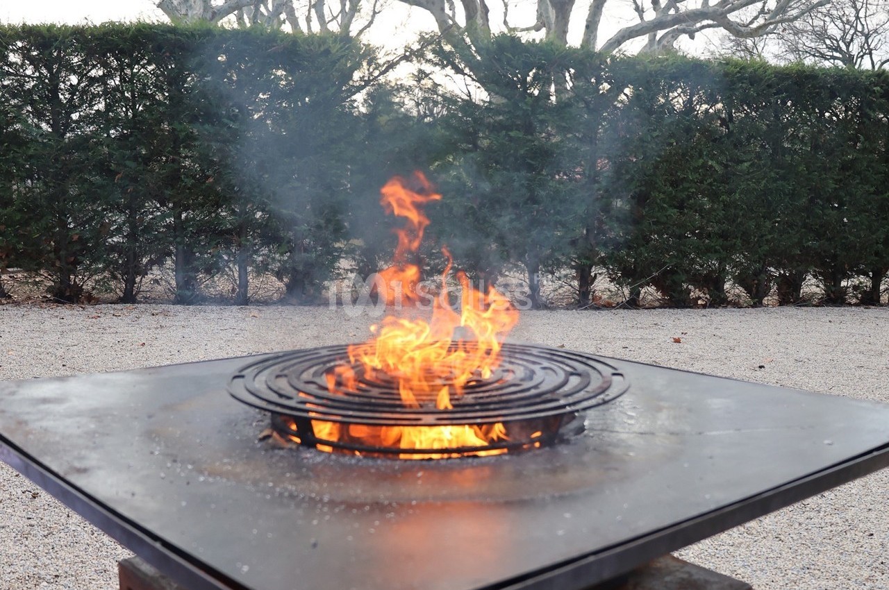 Brasero en métal avec grille circulaire, enflammé, installé sur un sol gravillonné devant une haie d'arbustes.