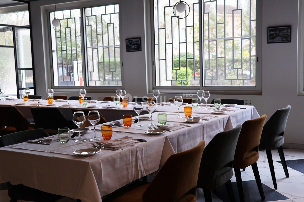 Salle de restaurant lumineuse avec tables dressées, nappes blanches, verres colorés et grandes fenêtres en arrière-plan.