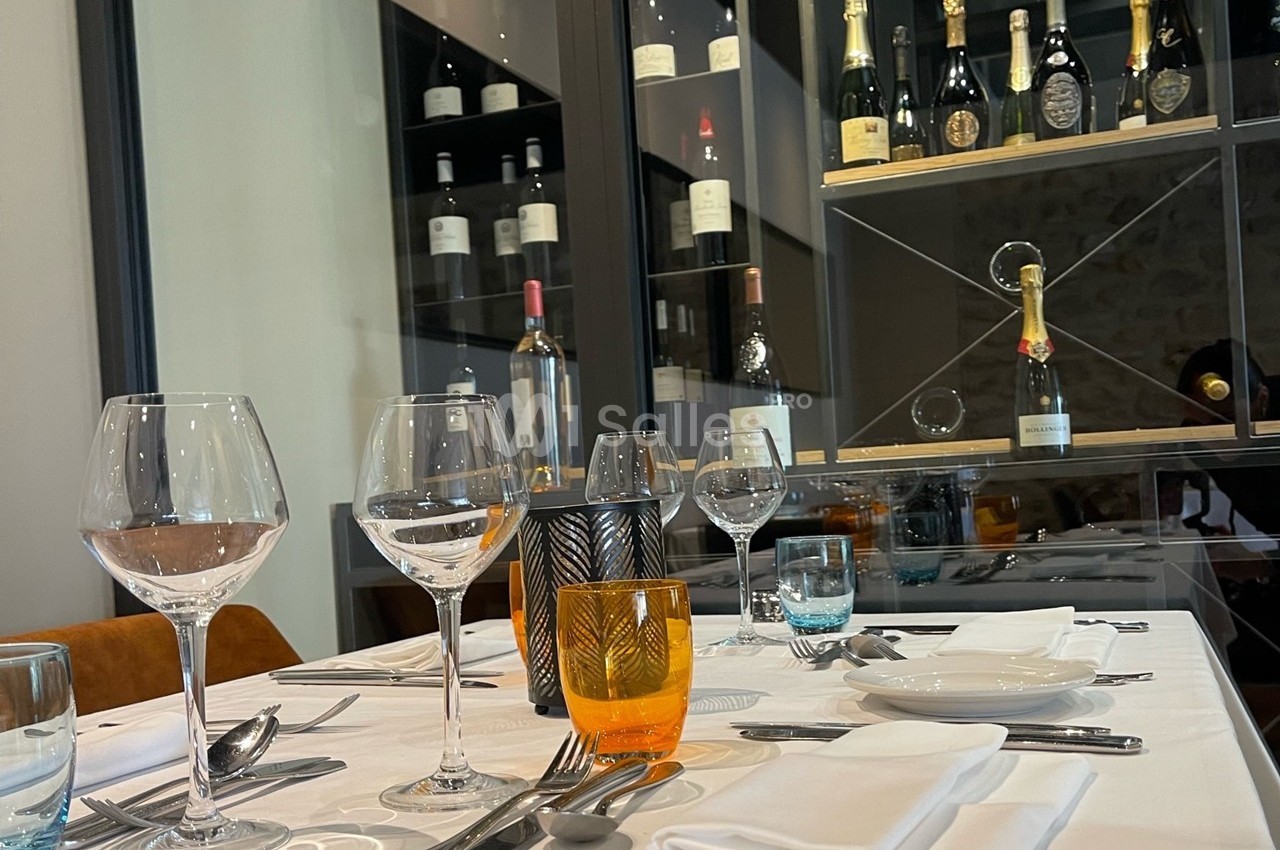 Table dressée avec verres, couverts et serviettes, devant une vitrine contenant des bouteilles de vin et de champagne.