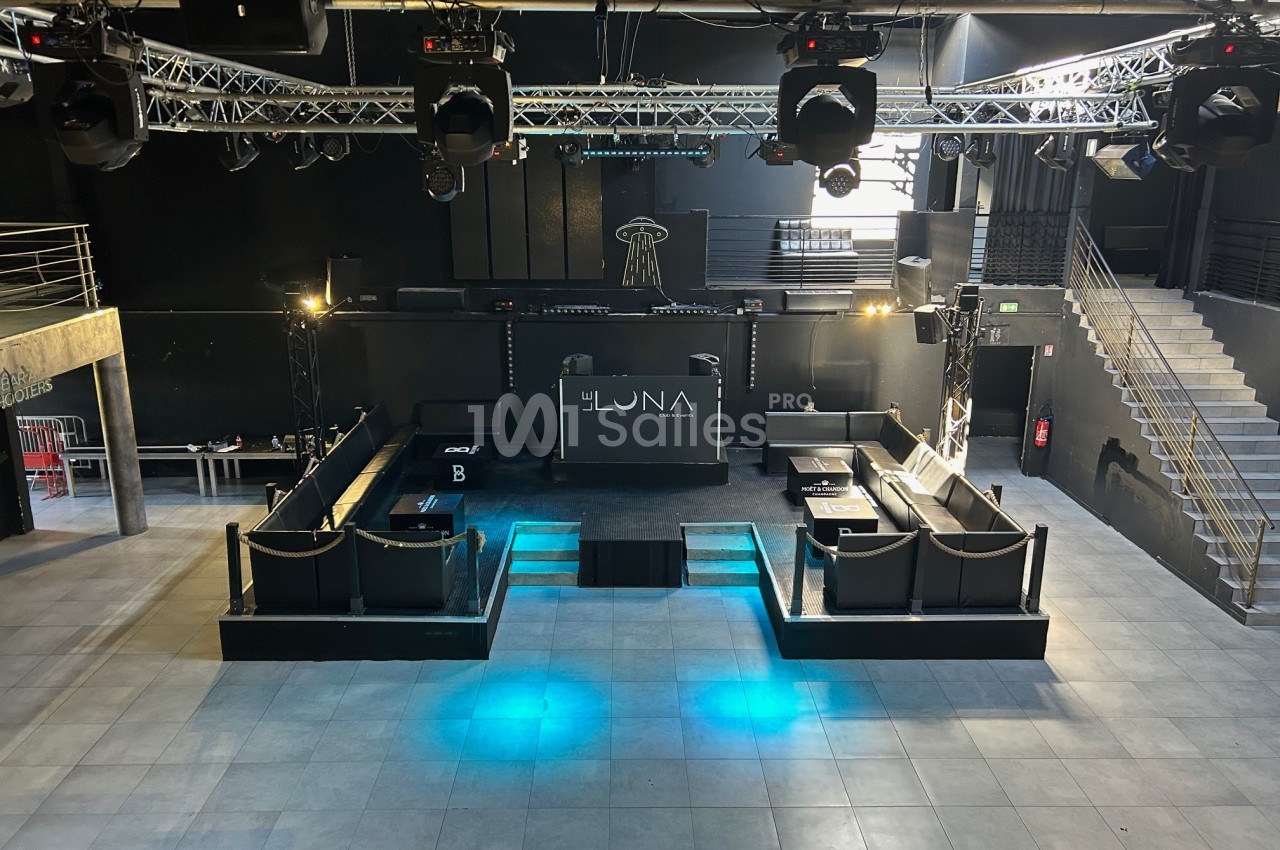Salle de concert vide avec scène centrale, éclairages suspendus, estrades et escaliers sur les côtés.