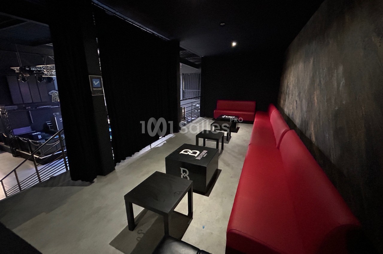 Espace lounge avec canapés rouges et noirs, tables basses et vue sur une scène dans une salle sombre.