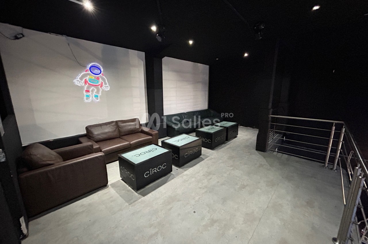 Espace lounge sombre avec canapés en cuir, tables basses marquées ’Cîroc’ et décoration lumineuse en forme d'astronaute.