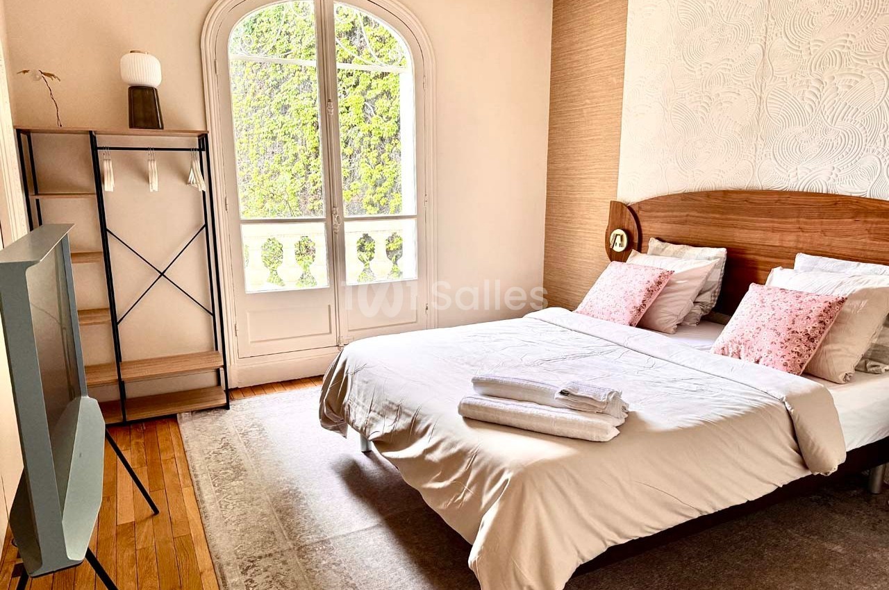 Chambre lumineuse avec lit double, étagère, télévision et grande fenêtre donnant sur des arbres.