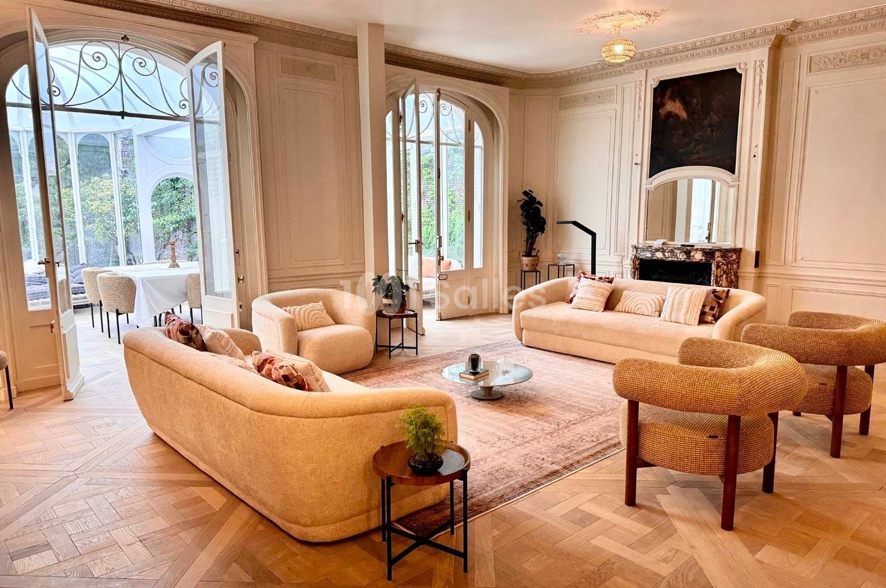 Salon lumineux avec parquet en chevrons, canapés beiges, table basse et grandes portes vitrées donnant sur un jardin.