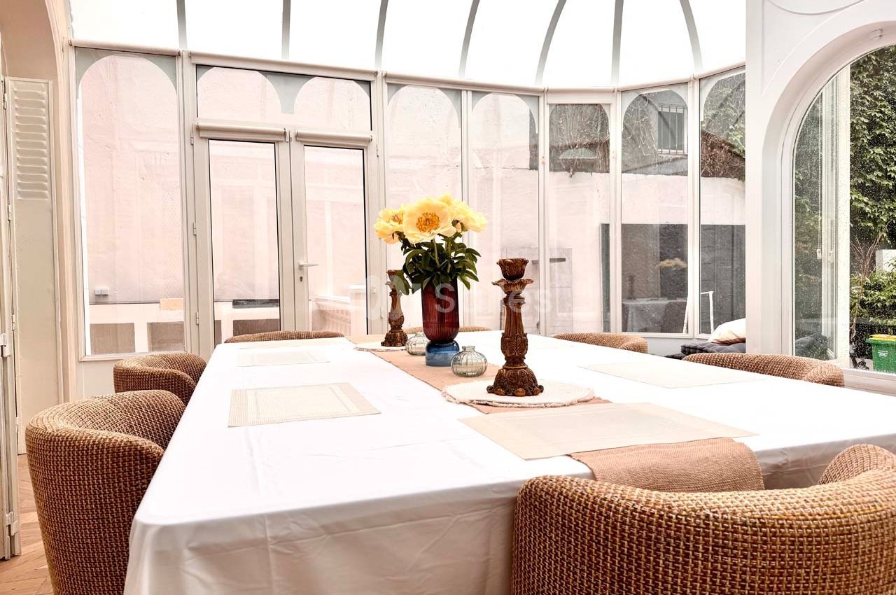 Salle à manger lumineuse avec une grande table blanche, chaises en rotin et un vase de fleurs jaunes au centre.