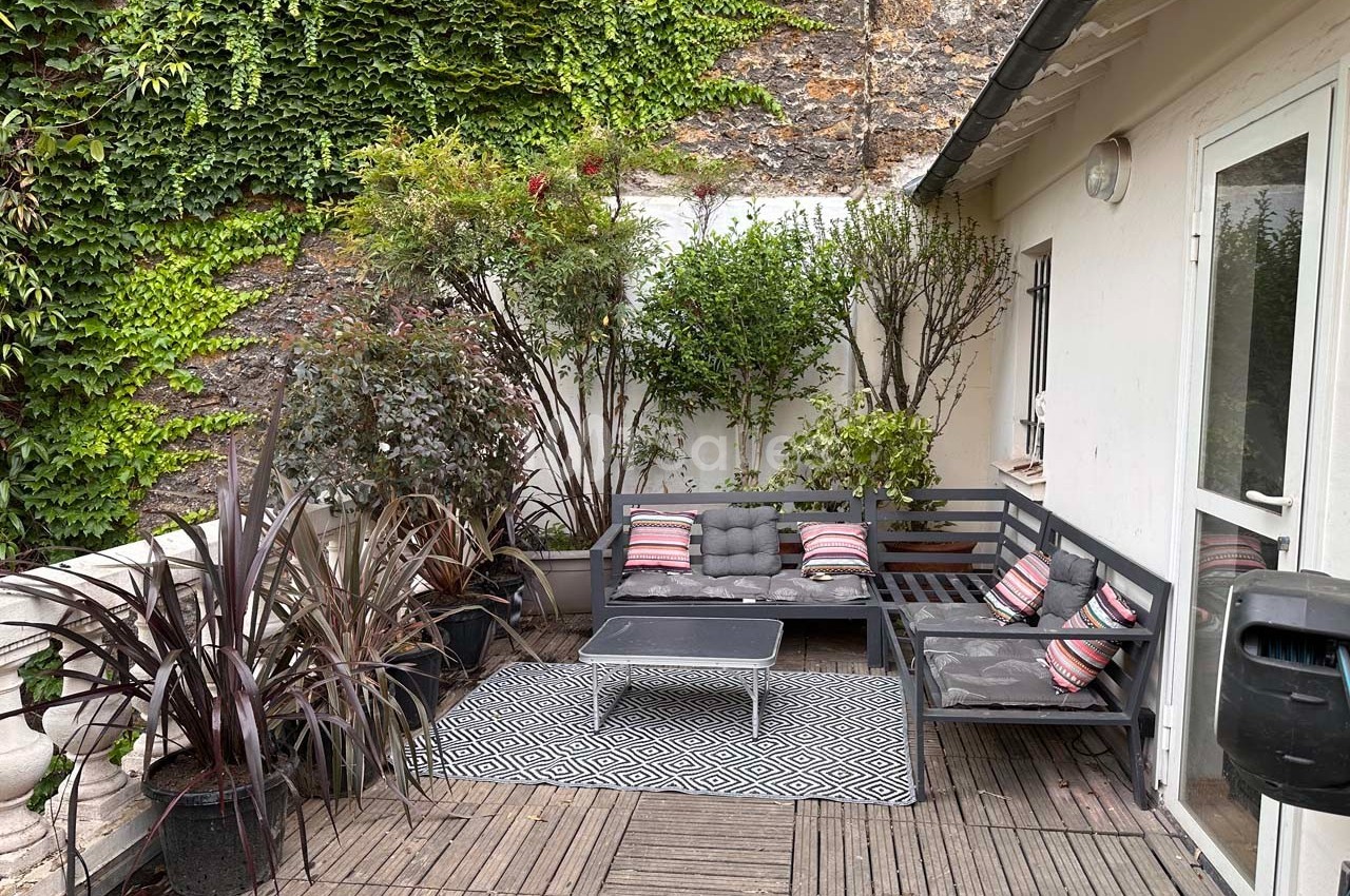Terrasse avec mobilier de jardin gris, coussins colorés, plantes en pots et mur recouvert de végétation.