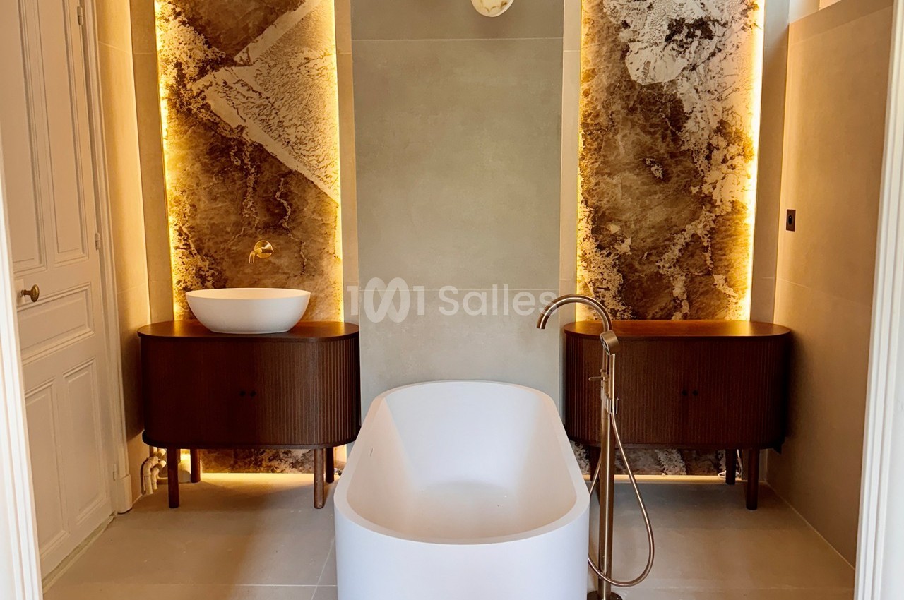 Salle de bain moderne avec baignoire centrale, deux vasques sur meubles en bois et éclairage mural chaleureux.