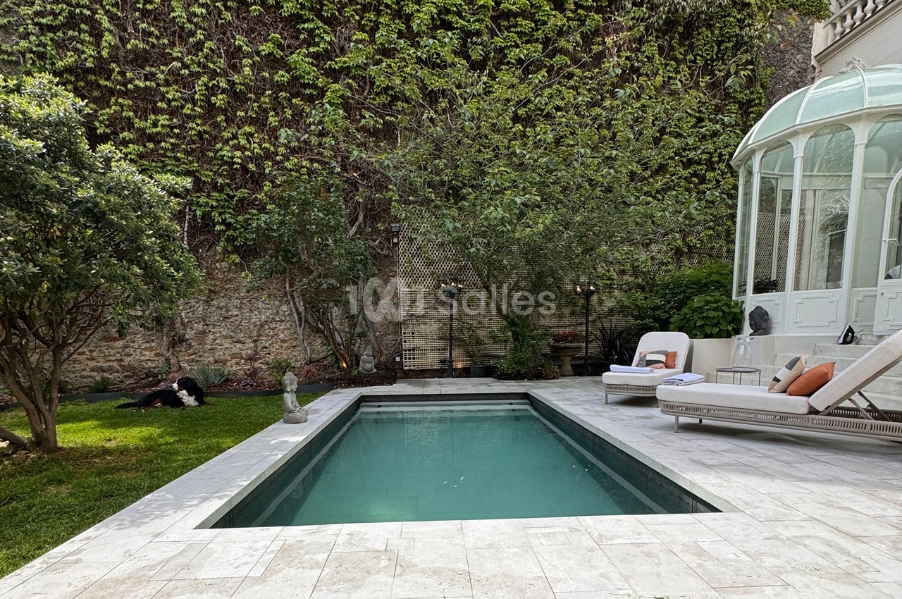 Piscine rectangulaire entourée de dalles claires, avec jardin verdoyant et véranda vitrée en arrière-plan.