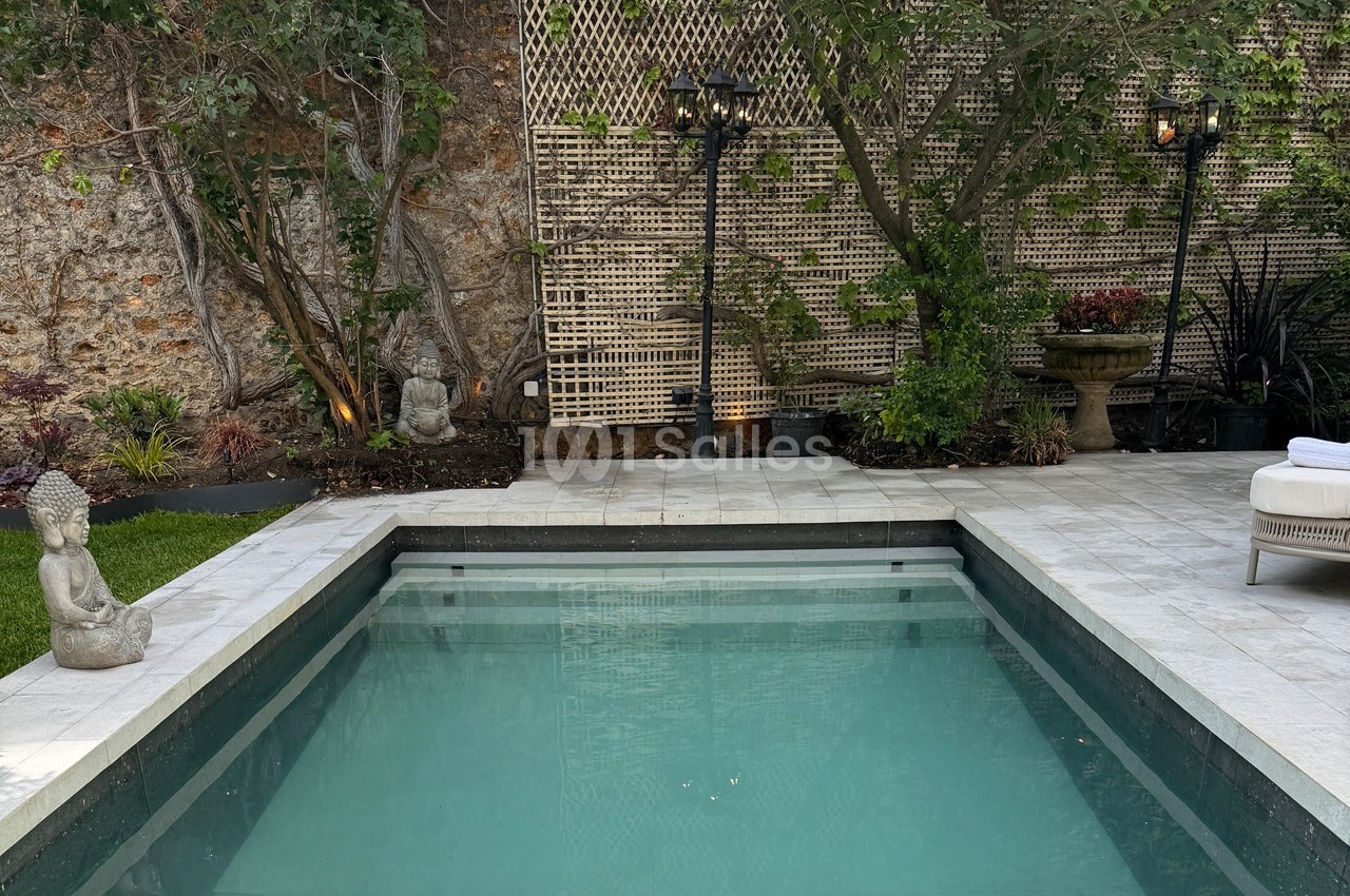 Piscine rectangulaire entourée de statues, végétation et éclairages, dans un jardin avec mur en pierre et treillis.