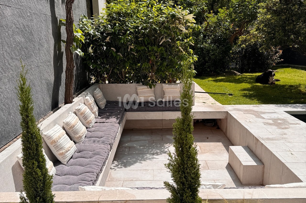 Coin salon extérieur encastré avec coussins gris, entouré de verdure et d'un jardin ensoleillé.