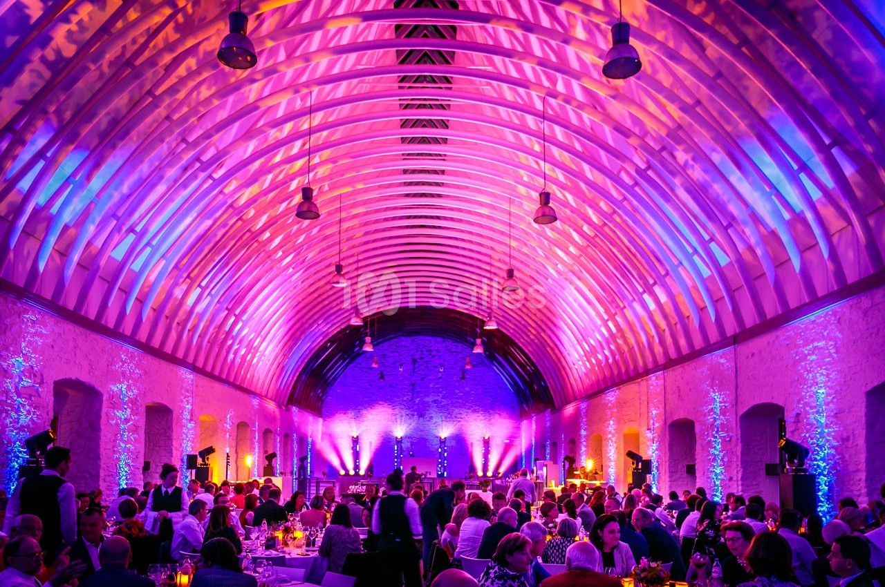 Salle voûtée illuminée par des lumières colorées, remplie de convives assis à des tables lors d'un événement.