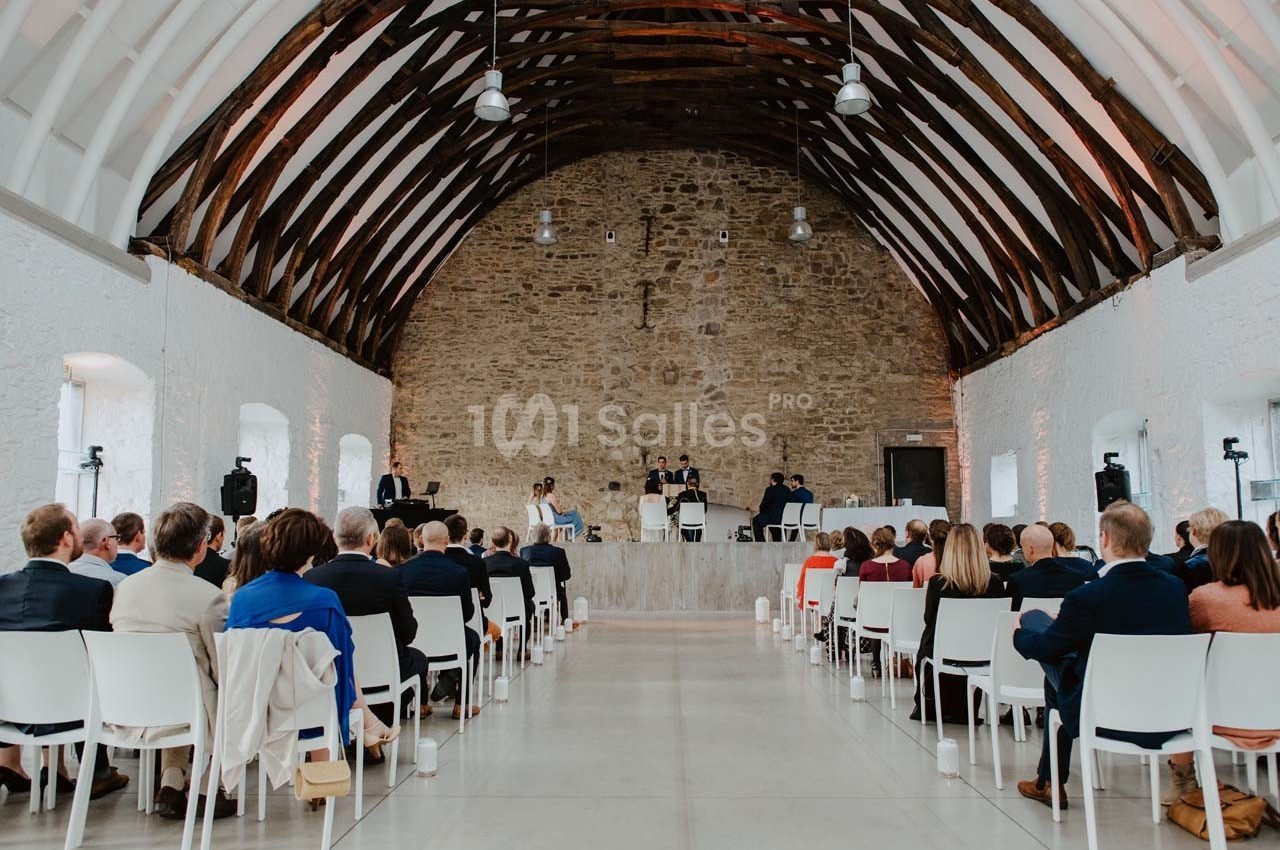 Salle avec des murs en pierre et poutres apparentes, aménagée pour une cérémonie avec des invités assis sur des chaises…