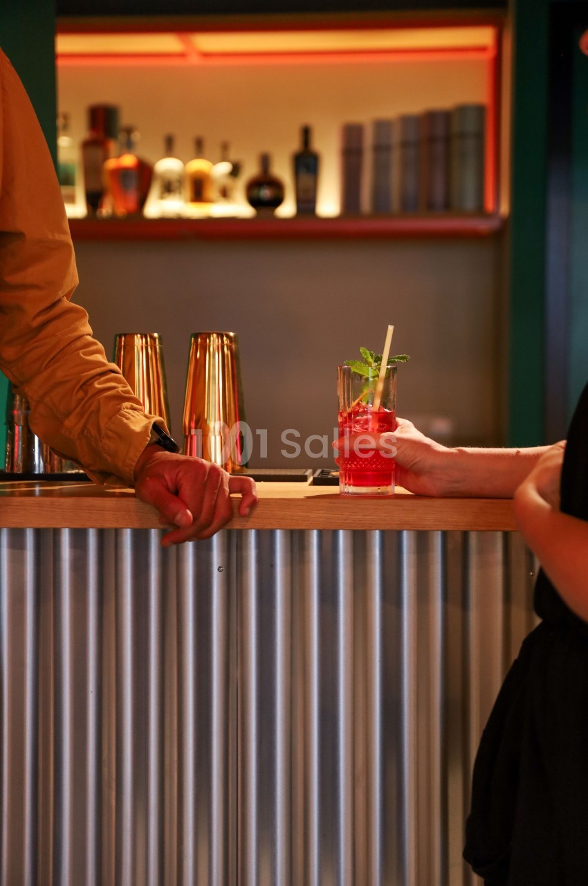 Un verre de cocktail rouge avec une paille et une feuille de menthe est tendu sur un comptoir de bar en métal.