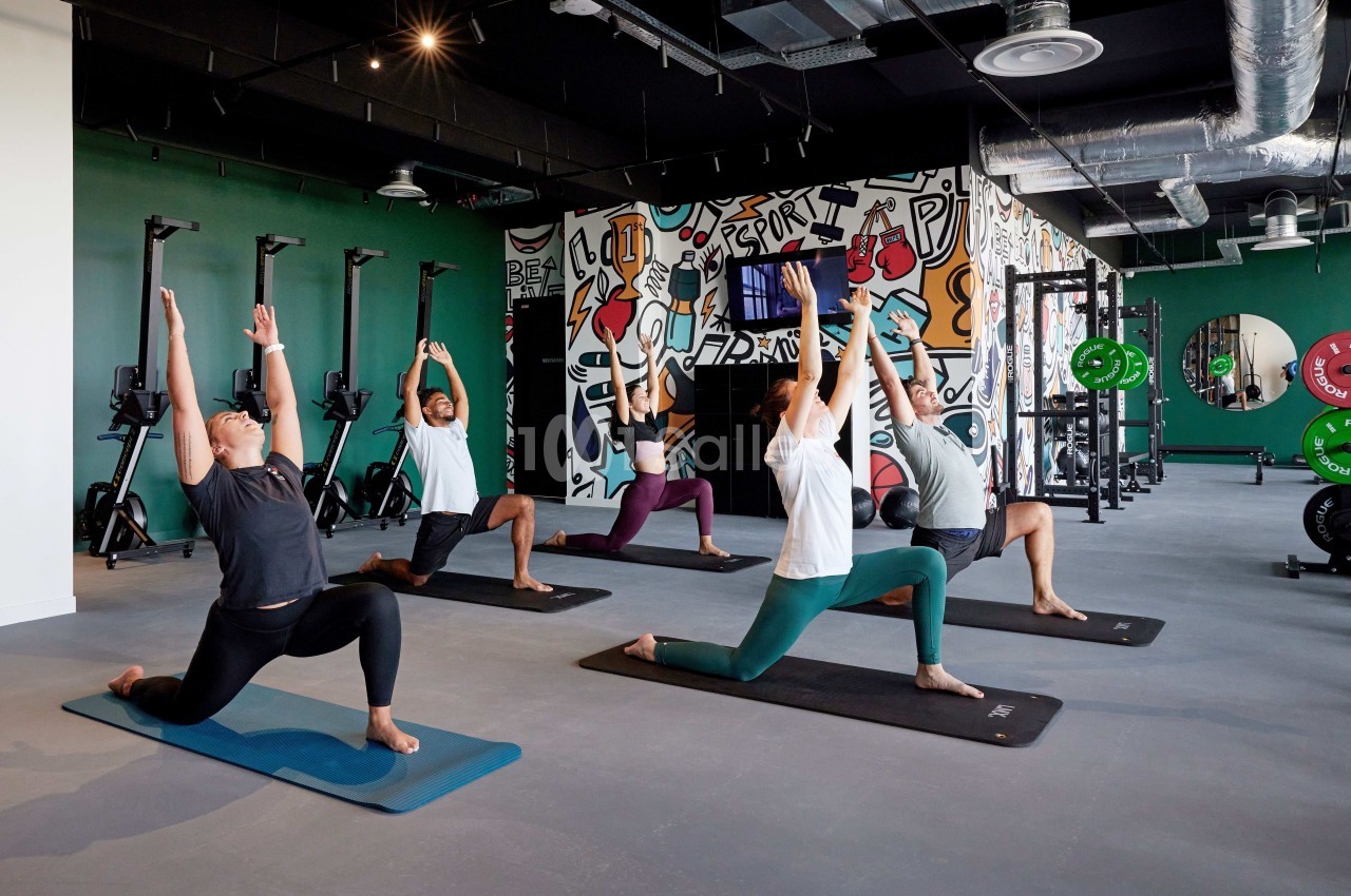Personnes pratiquant le yoga en groupe dans une salle de sport moderne avec des équipements et un mur décoré de graffitis.