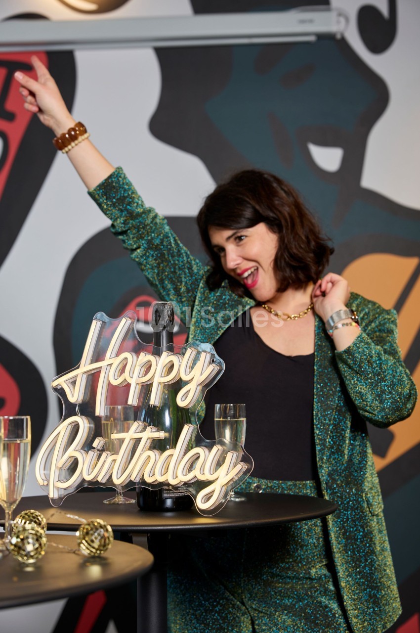 Femme souriante en tenue festive levant le bras près d'une table avec une décoration ’Happy Birthday’ et des verres.