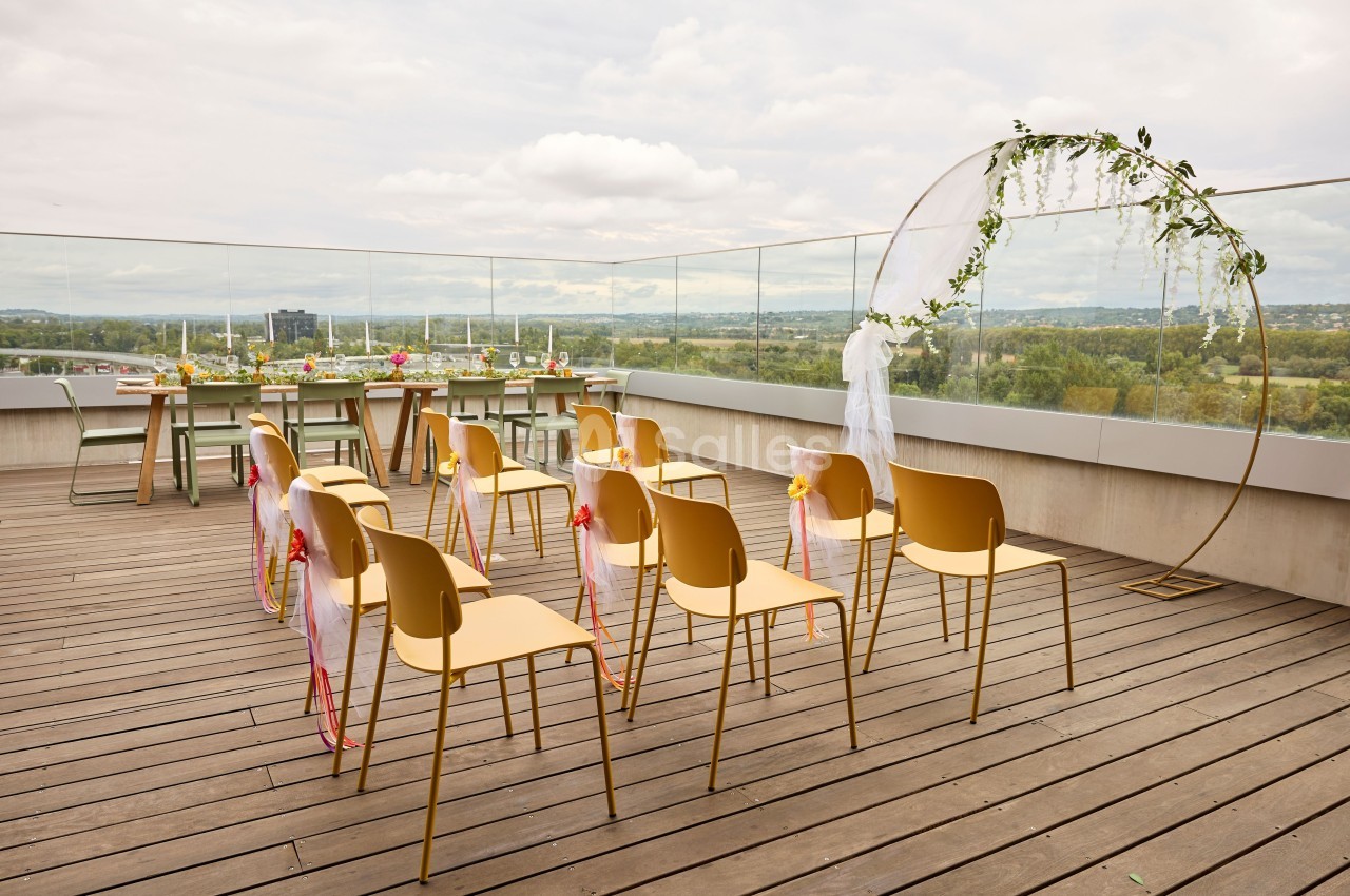 Chaises alignées sur une terrasse en bois avec arche décorée et vue dégagée sur un paysage verdoyant.