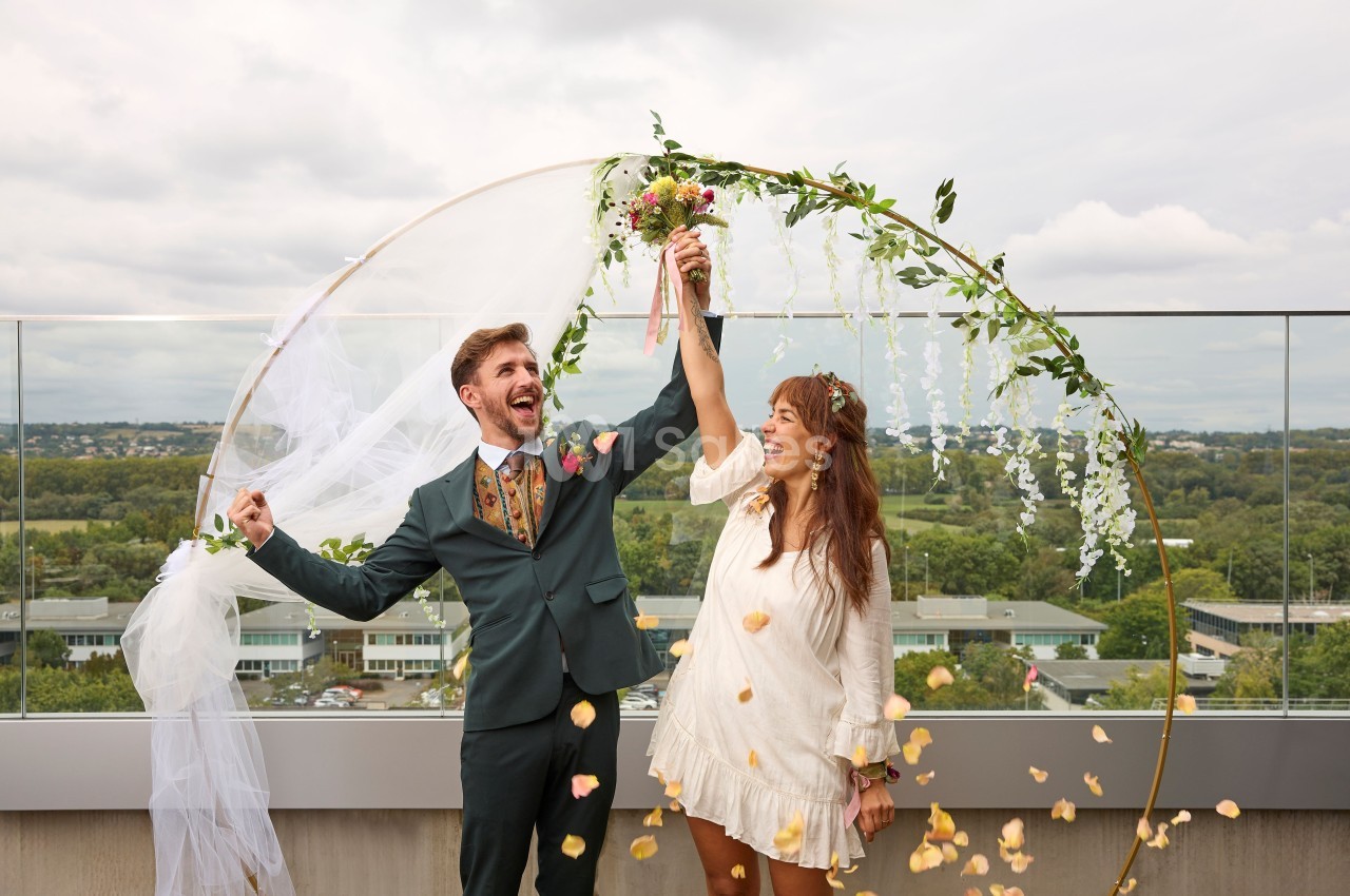 Un couple souriant sous une arche décorée de fleurs, tenant un bouquet, sur une terrasse avec vue dégagée.