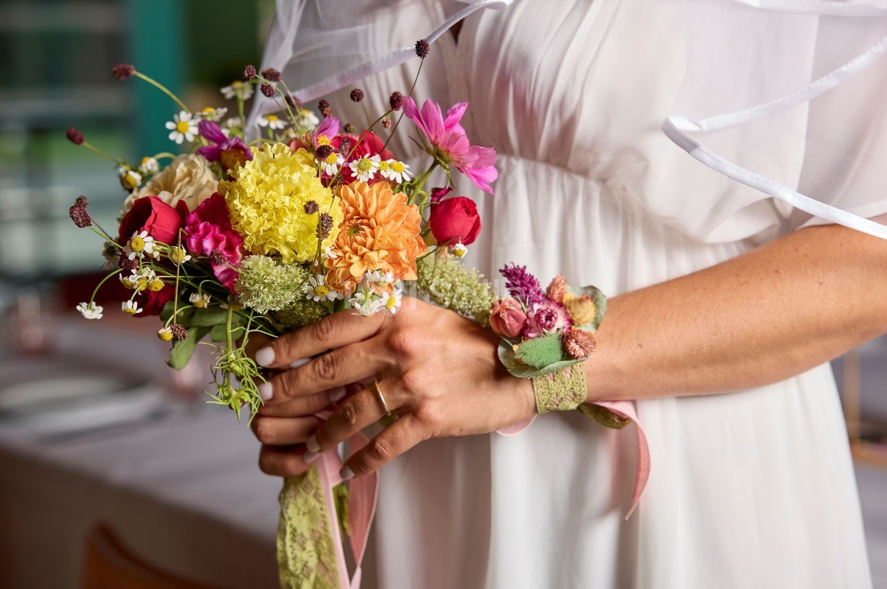 Une mariée en robe blanche tient un bouquet coloré de fleurs fraîches dans un décor intérieur lumineux.